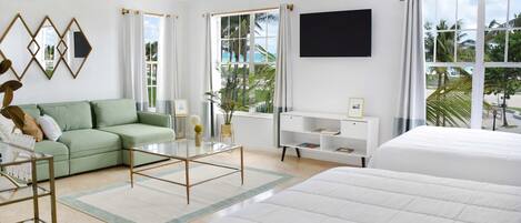 A modern hotel room with a green sofa, a glass coffee table, a white bed, and large windows offering a view of palm trees.
