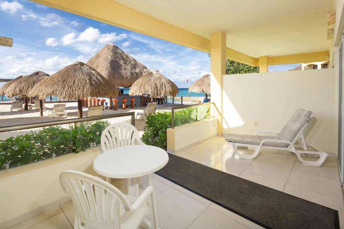 A balcony with white chairs and a table, overlooking a beach with thatched umbrellas and a clear blue sky.