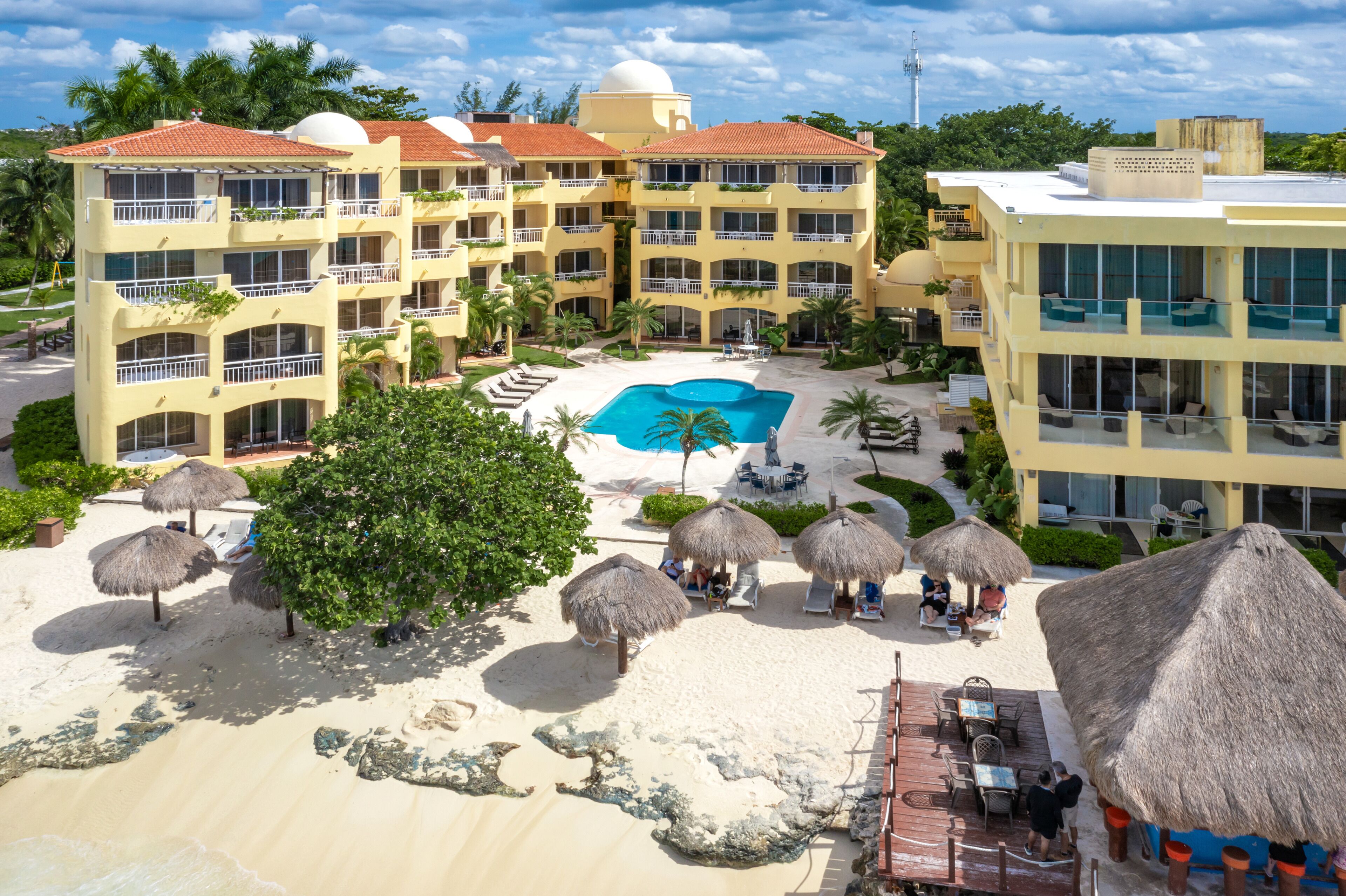 Un resort con varias edificaciones, una piscina central y sombrillas de techo de paja.