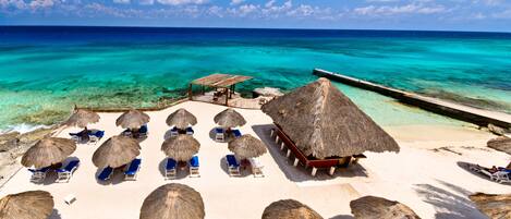 A beach with thatched-roof structures, lounge chairs, and a pier extending into the turquoise sea.