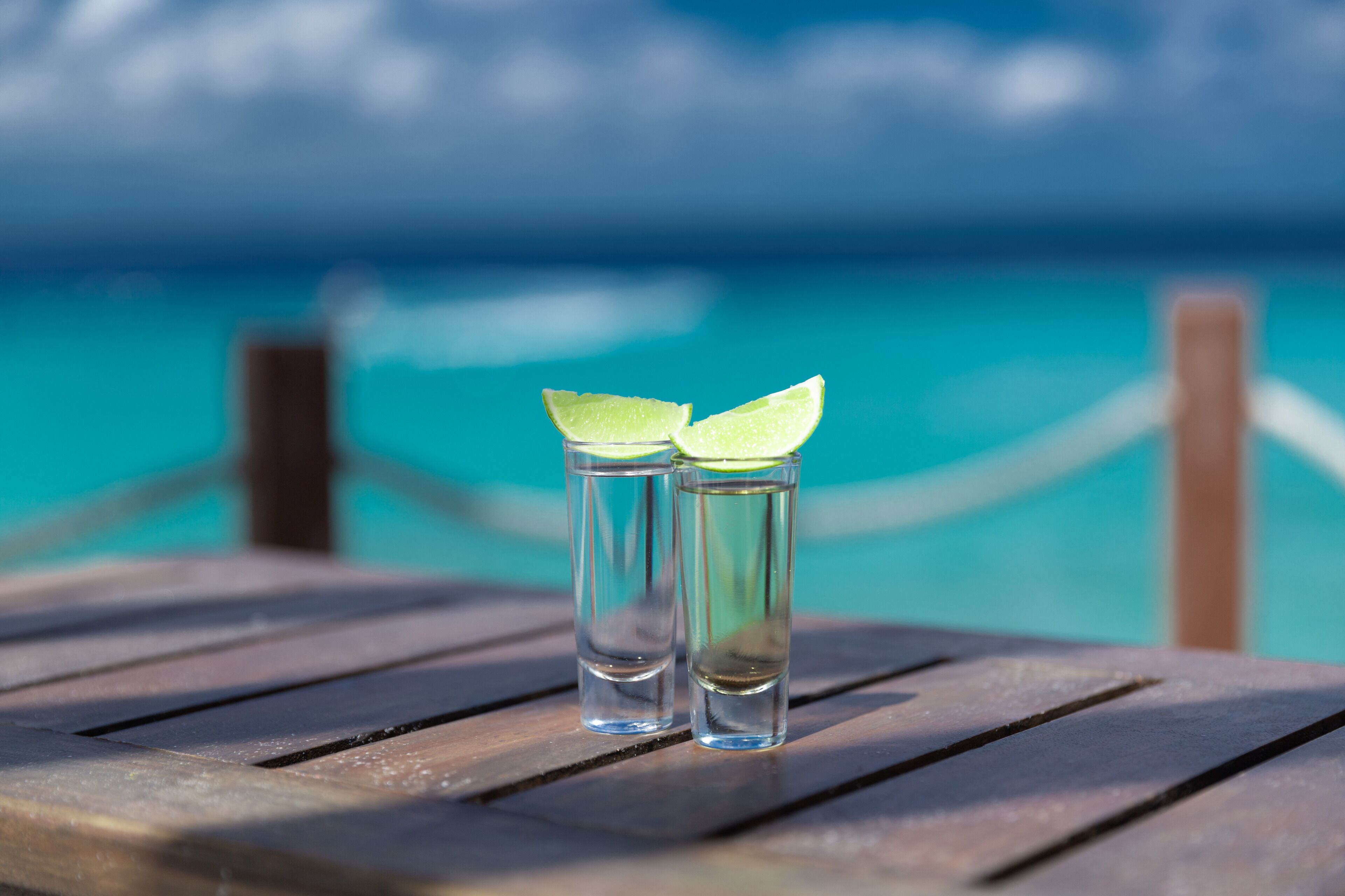 Two shot glasses with lime slices on a wooden table by a turquoise sea.