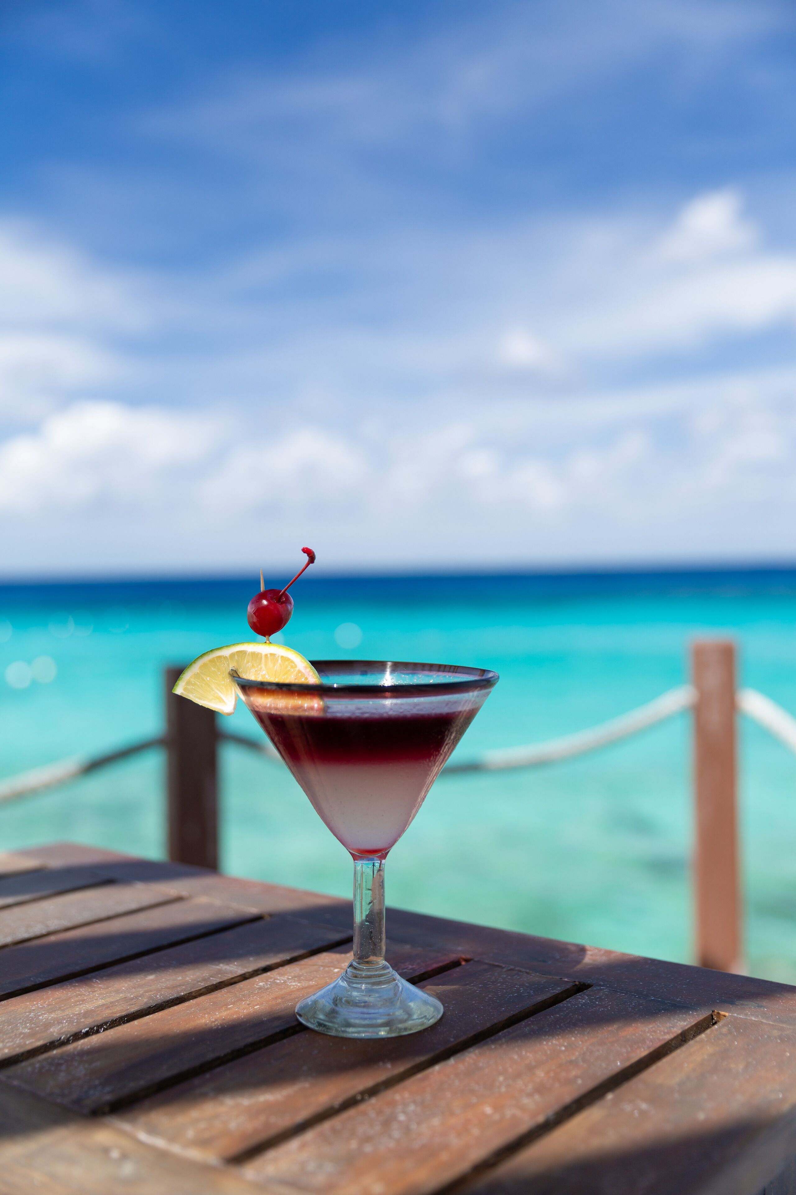 Un verre à martini, garni d’une cerise et d’une tranche de citron, est posé sur une table en bois, offrant une vue sur l’océan et un ciel bleu azur.