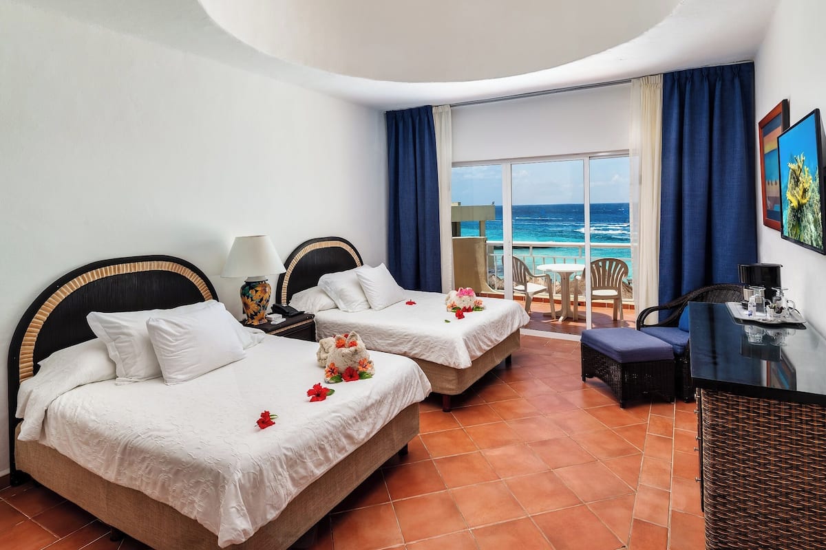 A hotel room with two beds, a desk, and a balcony with a view of the ocean.