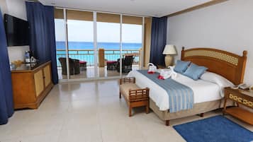 Habitación de hotel con cama, mesitas de noche, televisión, balcón con vista al mar y zona de estar con sillas.