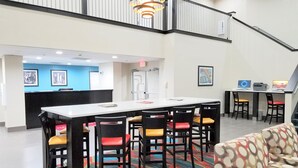 A hotel lobby with a central dining table, multiple chairs, a staircase, and a reception desk.