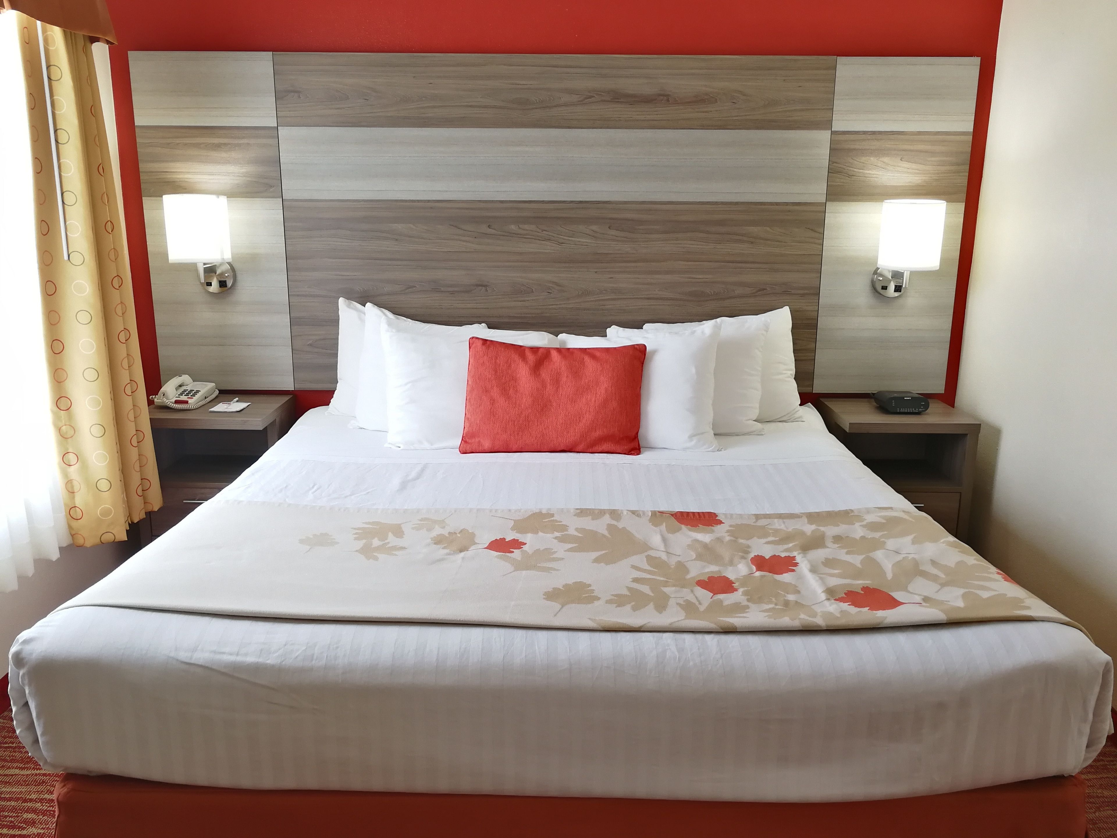 Une chambre d’hôtel avec un grand lit, du linge de lit blanc, un coussin rouge et deux tables de chevet équipées de lampes et d’un téléphone.