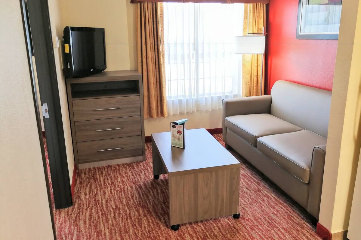 A hotel room with a sofa, a TV on a cabinet, a small table, and a window with curtains.