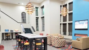 A modern hotel lobby with a large dining table, comfortable seating, a fireplace, and a TV screen displaying a sports game.