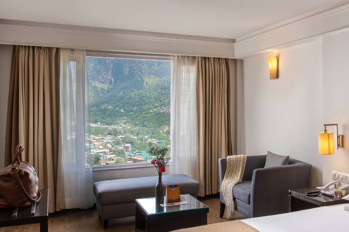 Habitación de hotel con un ventanal que ofrece vistas a un paisaje montañoso, un sofá, una mesita con un florero y una cama con ropa de cama impecable.