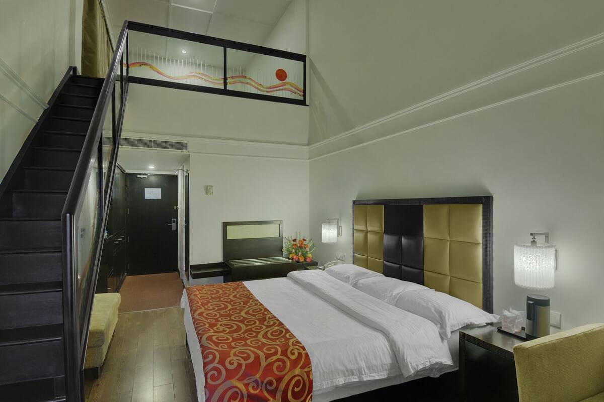 Una habitación de hotel con cama, mesita de noche, lámpara, escritorio, silla, sofá, escalera y puerta.