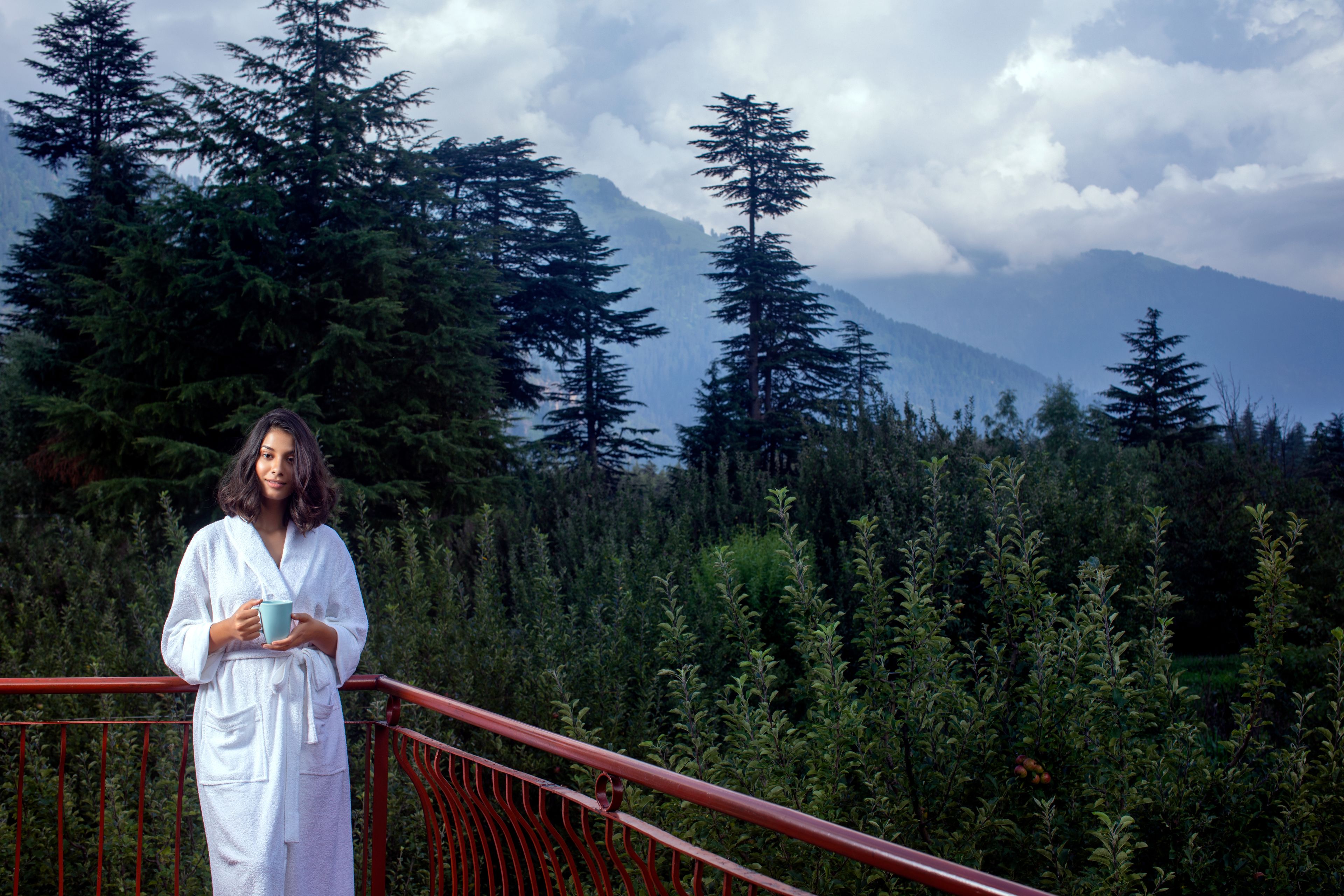 A person in a white robe standing on a red balcony with a view of a dense forest and mountains in the background.