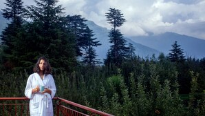 A person in a white robe standing on a red balcony with a view of a dense forest and mountains in the background.