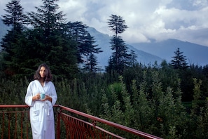 A person in a white robe standing on a red balcony with a view of a dense forest and mountains in the background. - Apple Country Resorts - A Vegetarian Place (Manali)