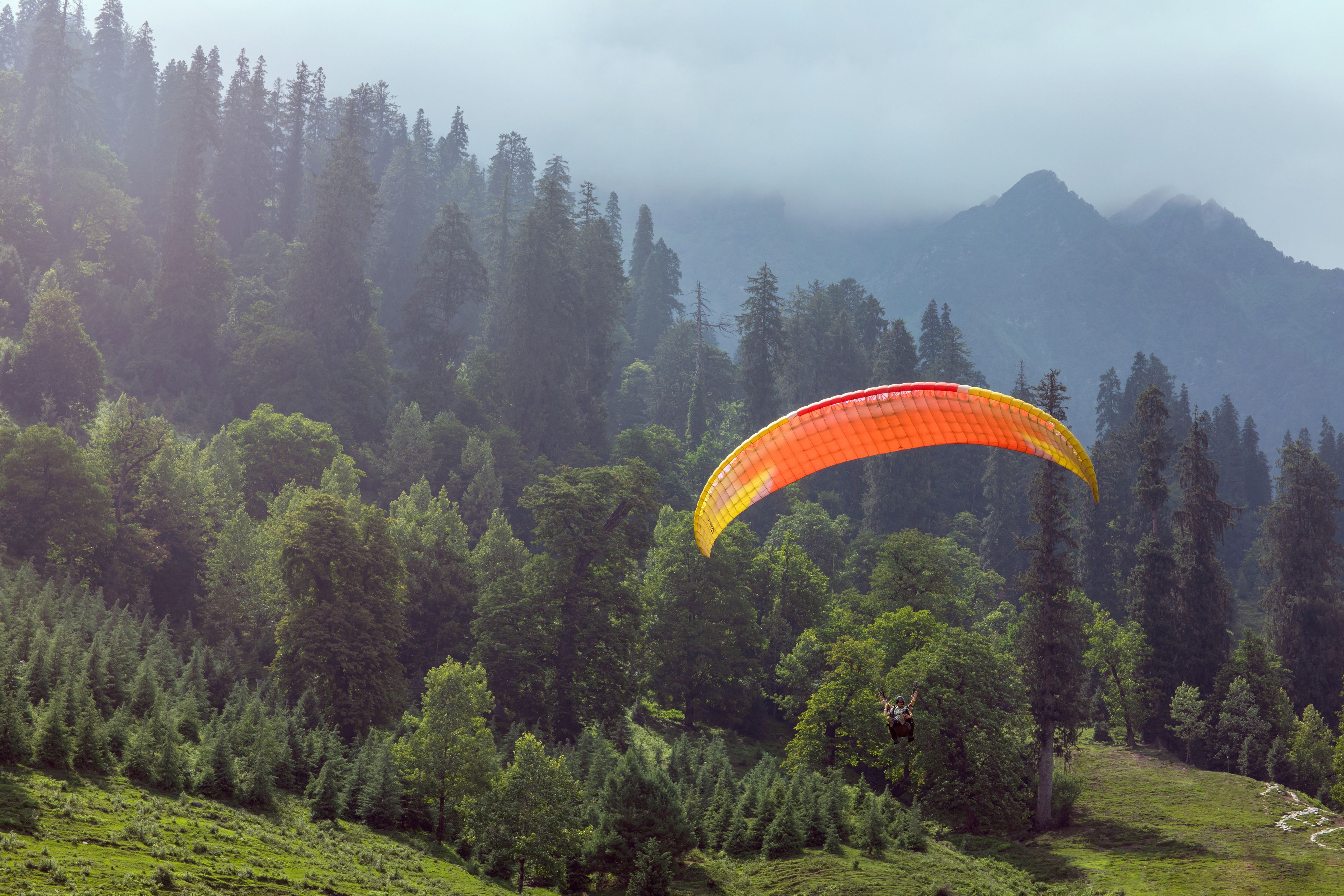 Une personne pratiquant le parapente au-dessus d’une zone montagneuse boisée.