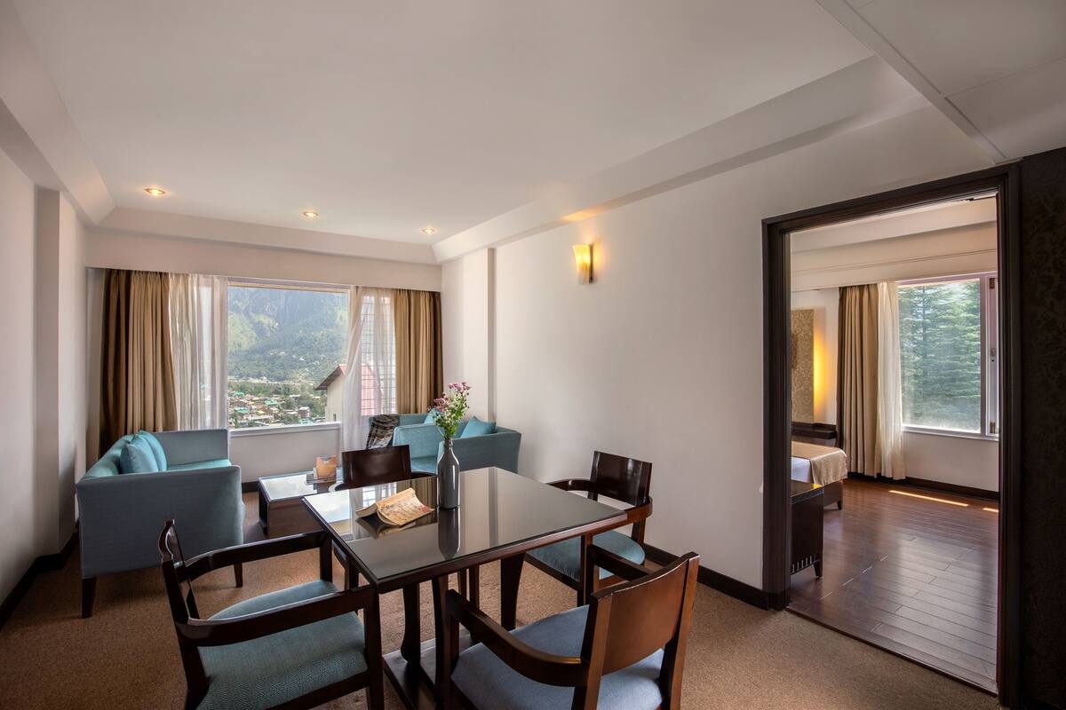 Habitación de hotel con una mesa de comedor, sillas, un sofá, una mesita con un florero y vistas al exterior.