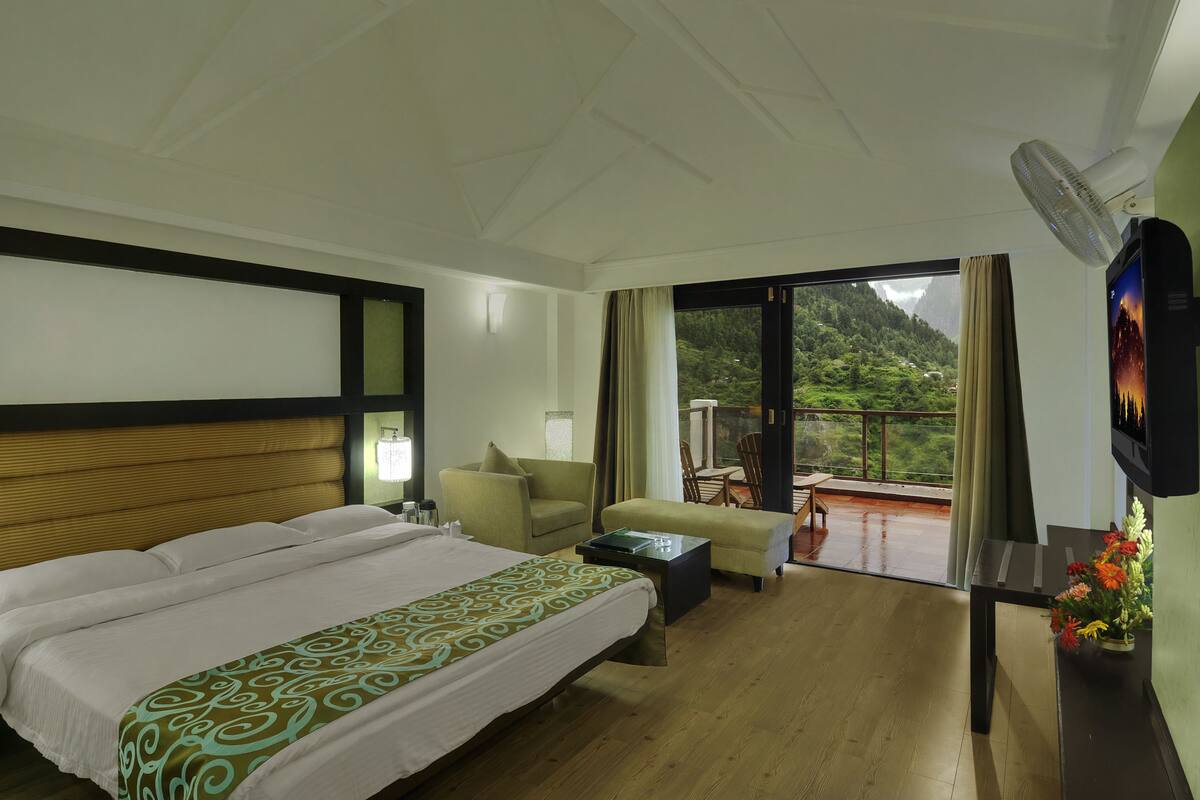 Habitación de hotel con una cama grande, un sofá, una mesita, un televisor y un balcón con vistas a la montaña.