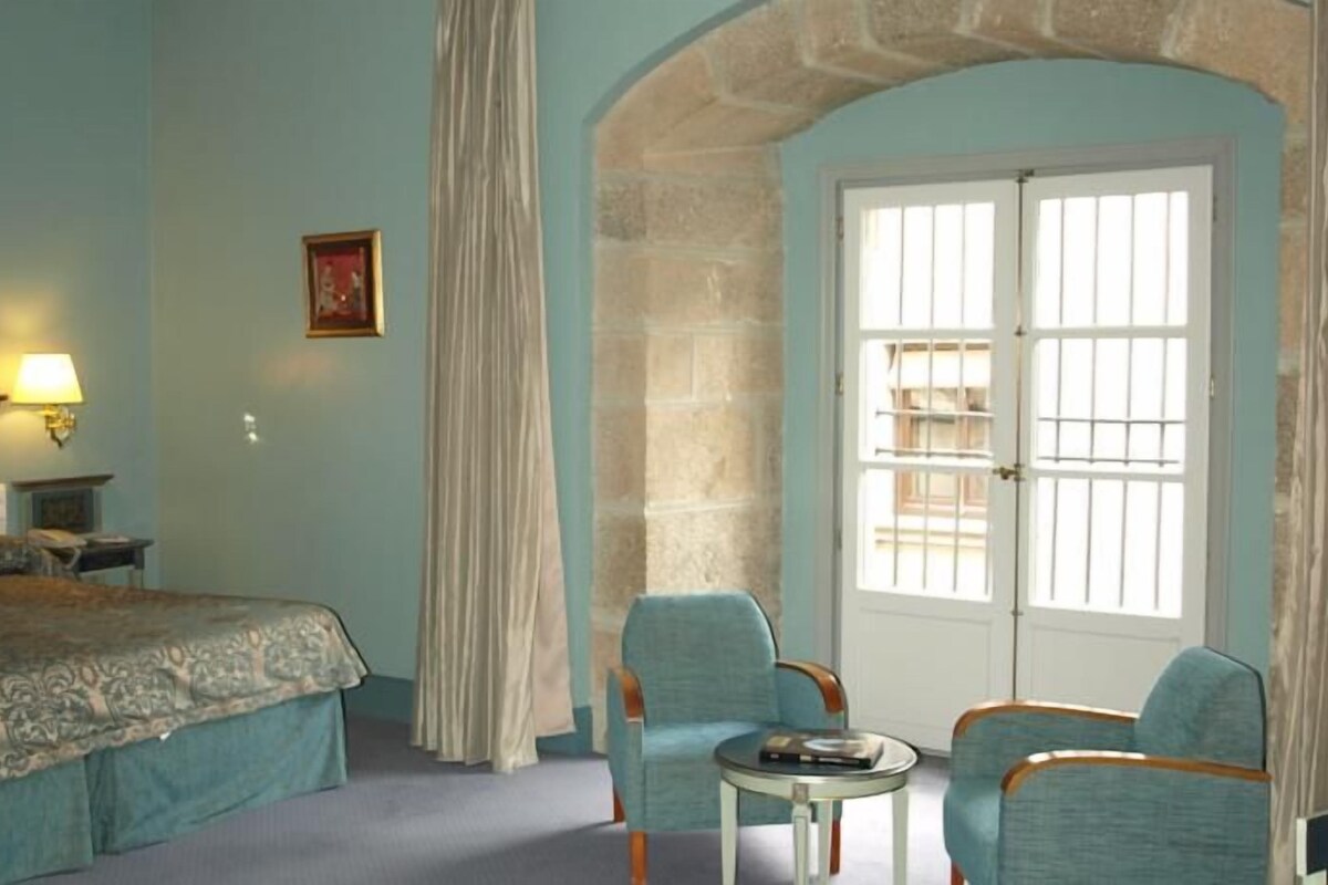 Habitación de hotel con cama, dos sillas, una mesita, lámpara, un cuadro y una ventana con cortinas.