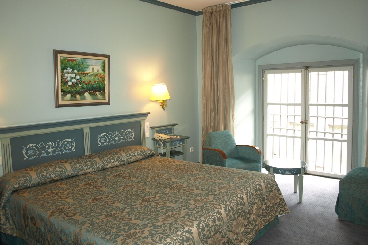 Habitación de hotel con una cama grande, una silla, una mesita, una lámpara y una puerta con vistas.