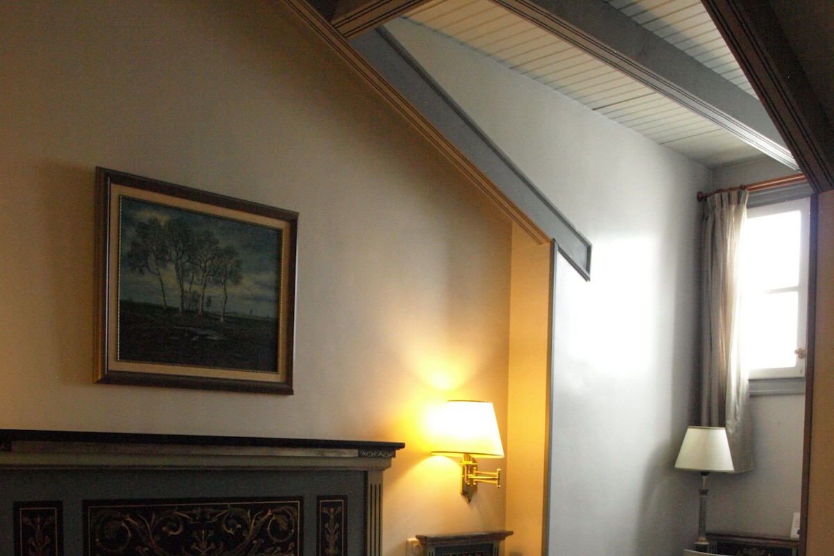 A hotel room with a large bed, a painting on the wall, a lamp, and a desk with a chair.