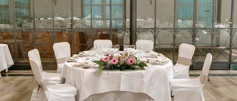 Una mesa redonda para seis personas, con sillas blancas, mantel blanco y un centro de mesa con flores.