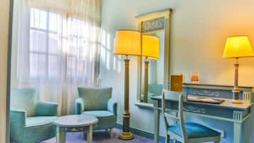 A hotel room with two armchairs, a small round table, a desk with a lamp, a mirror, and a window with sheer curtains.