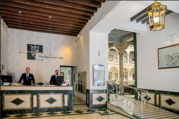 A hotel reception area with a marble counter, two receptionists, a staircase, and a view of the hotel's interior. - Hotel Palacio de los Velada (Ávila)