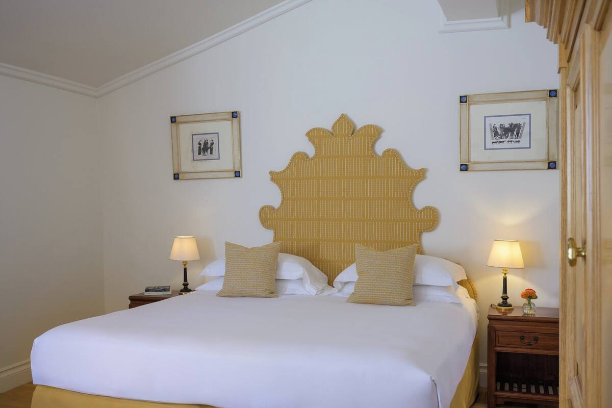 A neatly made bed with white linens and two pillows, a wooden headboard, two bedside tables with lamps, and framed pictures on the walls.