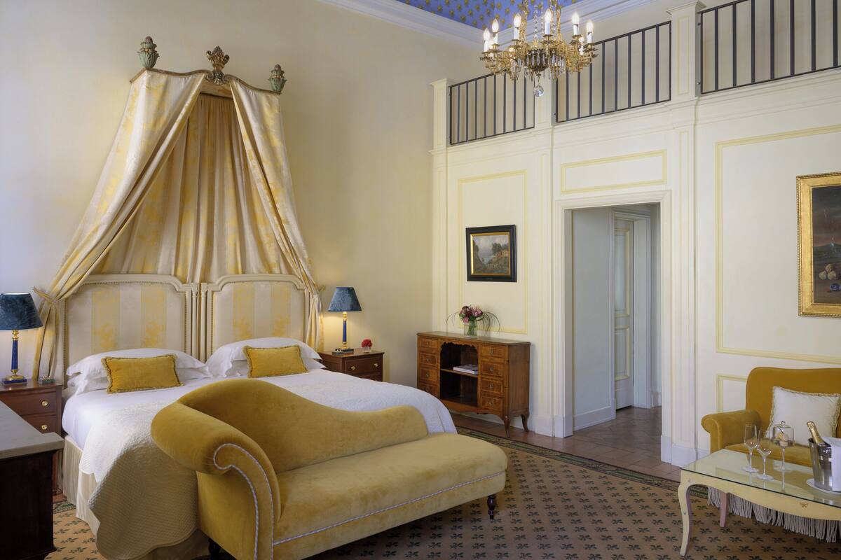 A luxurious bedroom with a four-poster bed, a chaise lounge, a nightstand with lamps, a wooden dresser, and a glass coffee table with wine glasses.