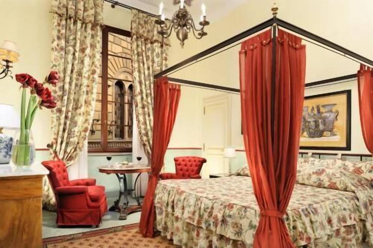 A four-poster bed with floral bedding, a red chair, a small table, and a chandelier.