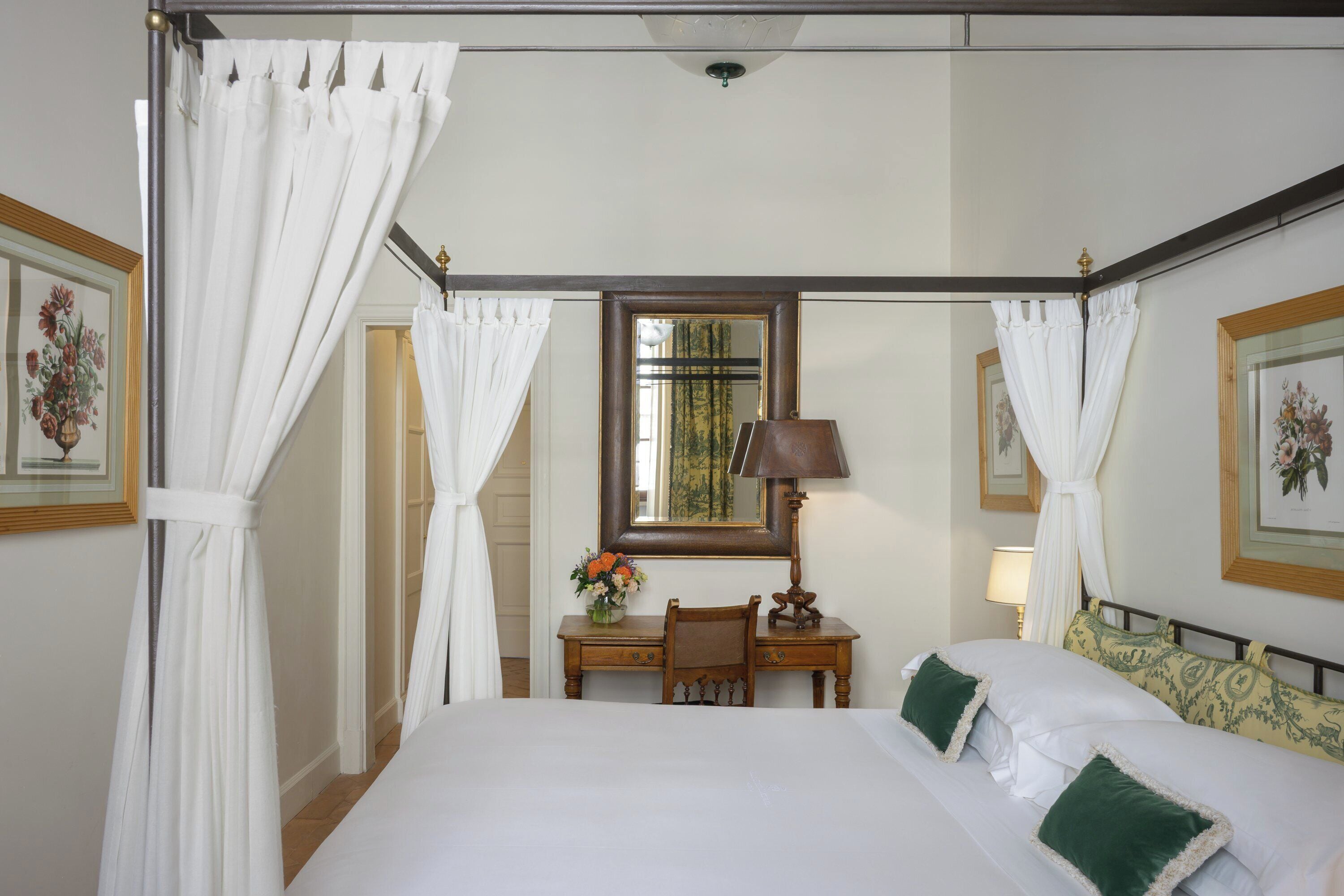 Un lit à baldaquin avec du linge de lit blanc et des coussins verts, un bureau en bois avec une lampe et un miroir au-dessus.