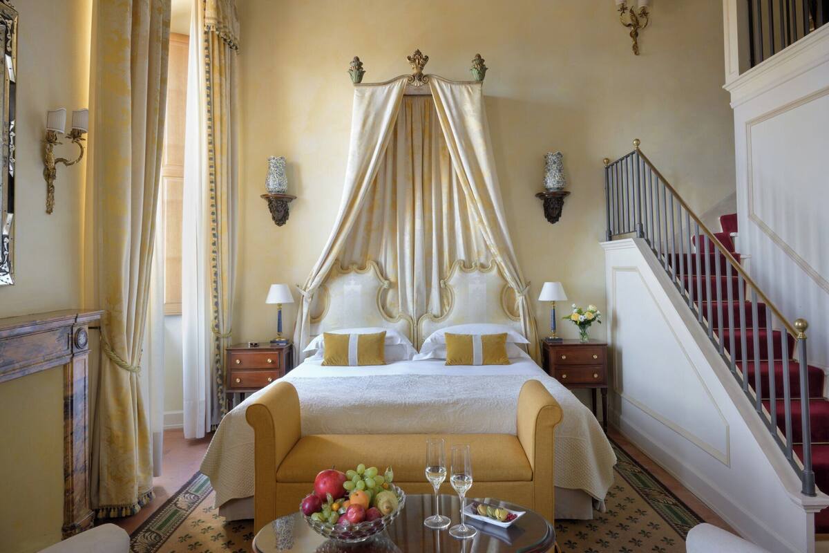 A luxurious bedroom with a four-poster bed, a sitting area with a sofa, a dining table with fruit and glasses, and a staircase.