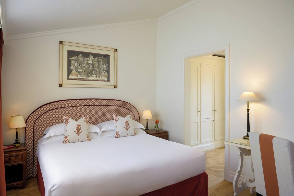 A neatly made bed with white linens and patterned pillows, a framed artwork above the bed, a bedside table with a lamp, and a chair with a cushion.