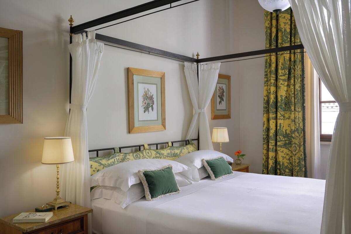 A neatly made bed with white linens and green pillows, a bedside table with a lamp, and a framed picture on the wall.