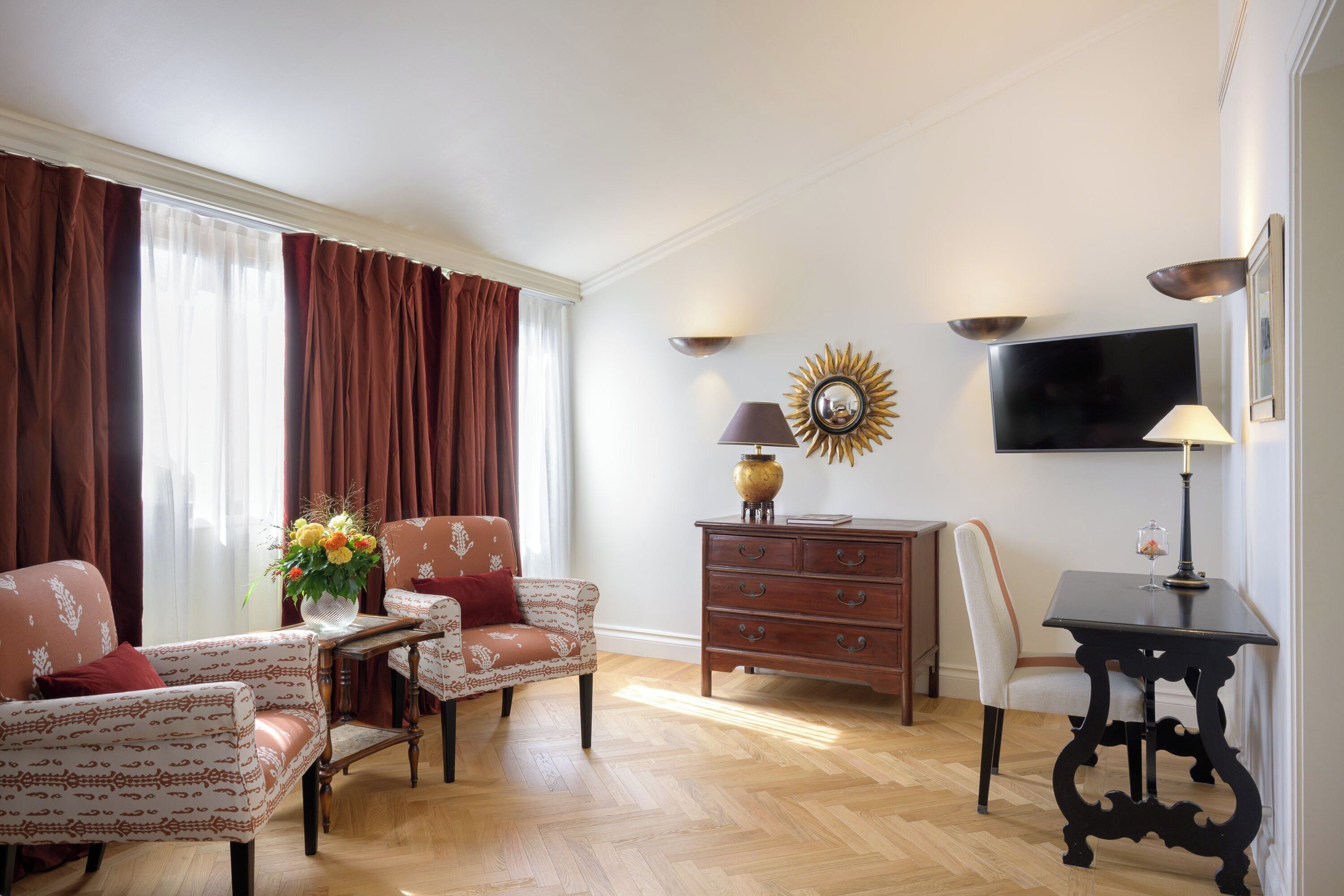 A living room with a sofa, armchair, and a small table with a lamp. There is a TV mounted on the wall and a decorative mirror.