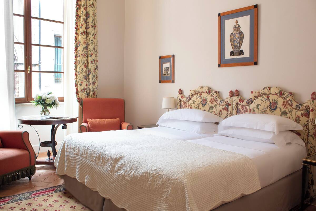 A hotel room with a bed, a chair, a table with a vase, and two framed pictures on the wall.