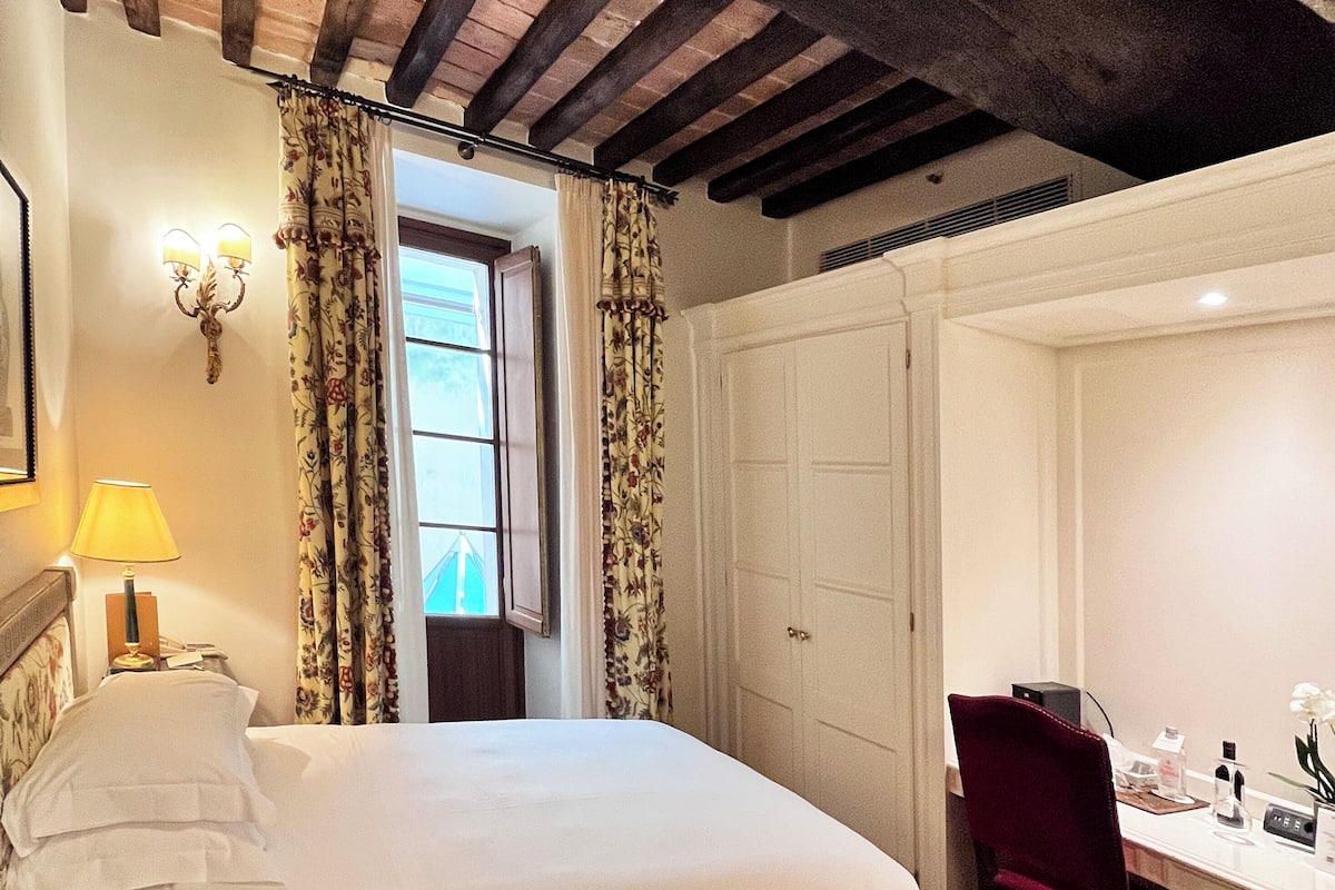 A bedroom with a bed, a desk, and a chair. There is a window with curtains, a lamp, and a wall-mounted light fixture.