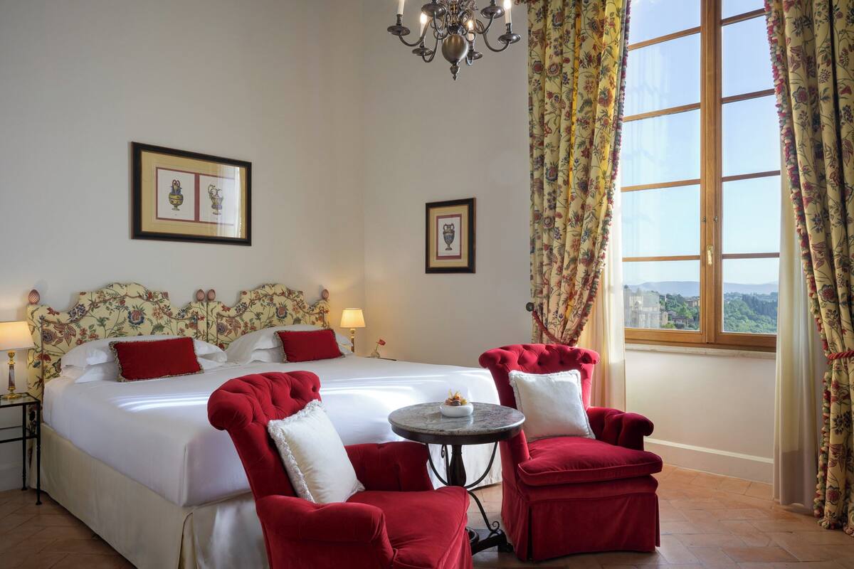 A hotel room with a large bed, two red armchairs, a small round table, a chandelier, and a window with curtains.