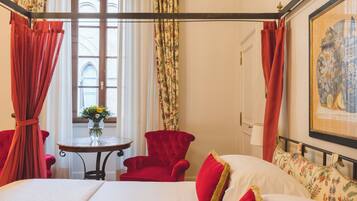 Quarto de hotel com cama, poltronas vermelhas, mesa pequena com um vaso de flores e janela com cortinas.