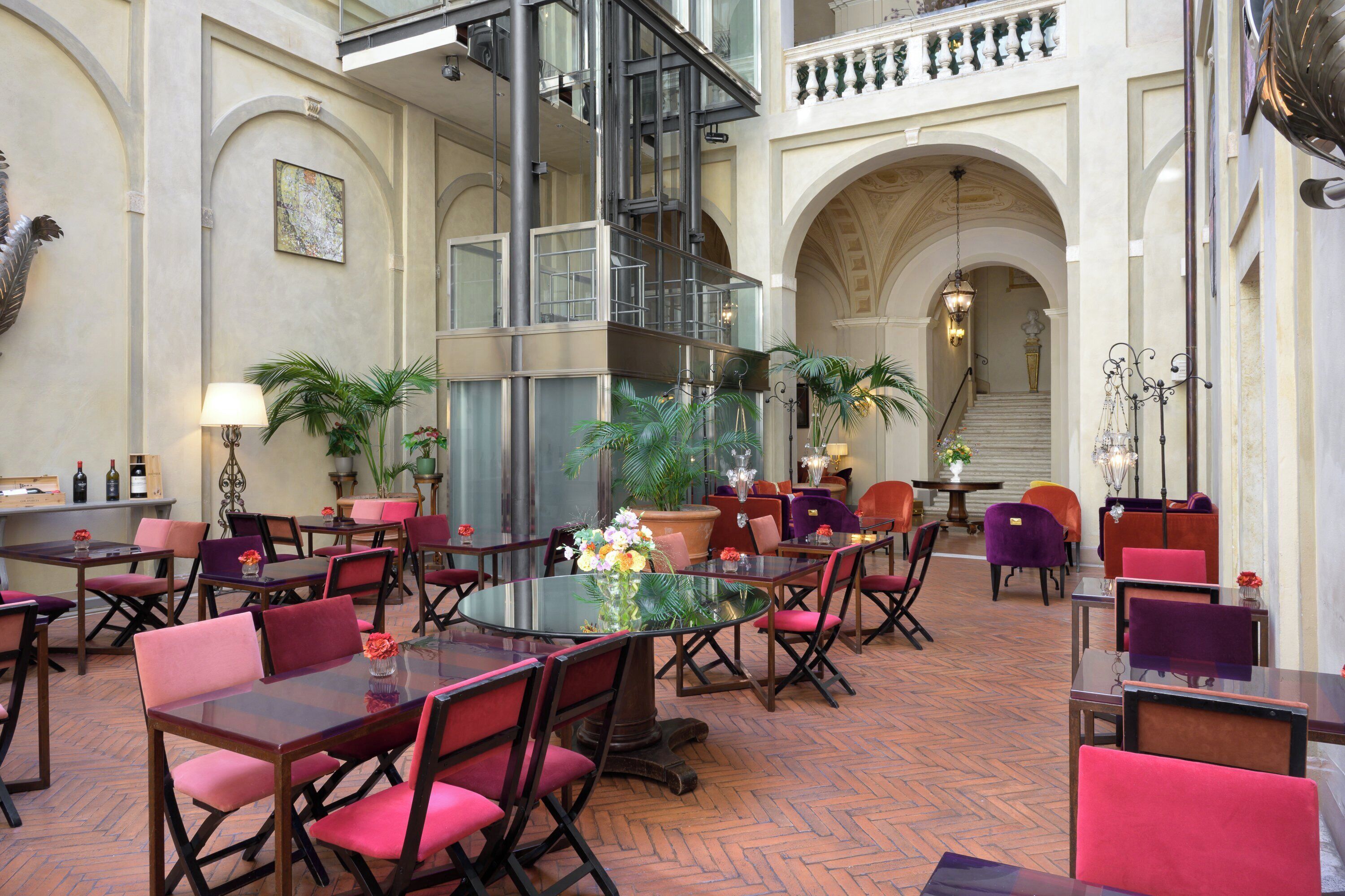 A spacious lobby with multiple dining tables and chairs, a central staircase, and decorative plants.