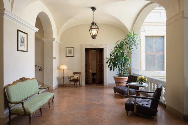 A spacious hallway with a green upholstered bench, a wooden chair, a table with a potted plant, and a large window with a decorative door. - Grand Hotel Continental Siena – Starhotels Collezione (Siena)
