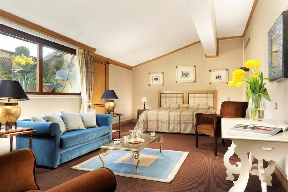 A room with a blue sofa, a bed with a beige bedspread, a white table, and a window with curtains.