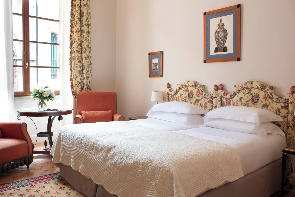 A hotel room with a bed, a chair, a table with a vase, and two framed pictures on the wall.