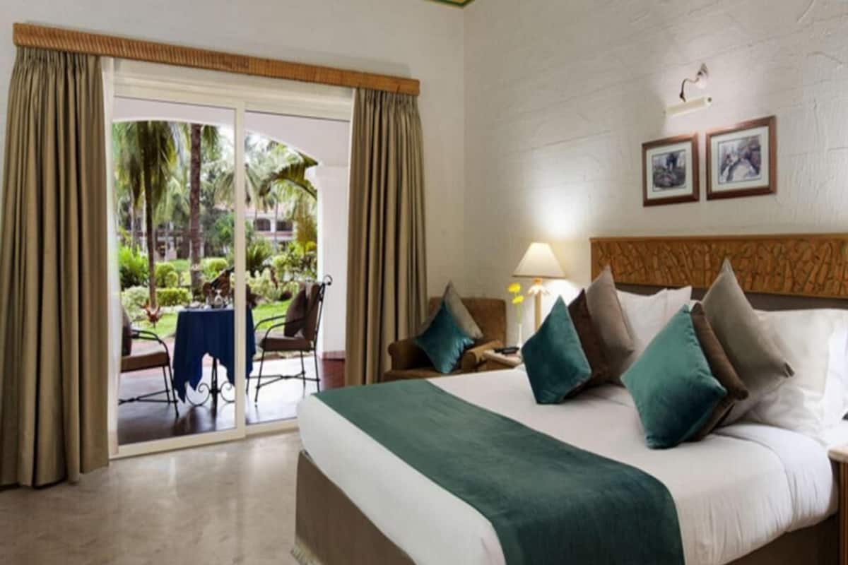 A hotel room with a large bed, a view of a garden through a sliding door, a table with chairs, and a lamp on a bedside table.