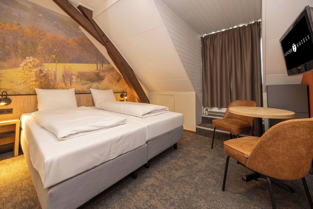 Habitación de hotel con cama, dos sillas, una mesita, televisor y un cuadro en la pared.
