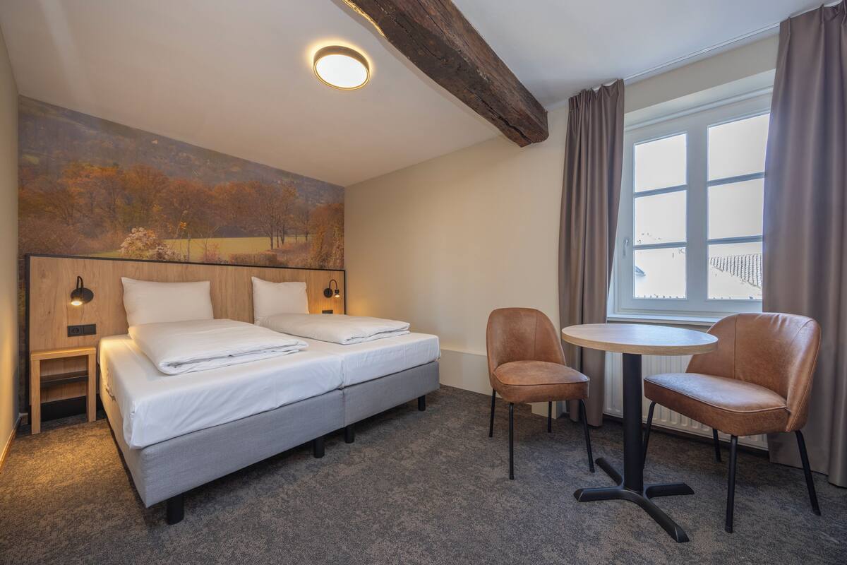 Habitación de hotel con cama doble, una mesita con dos sillas, lámpara de pared y ventana con cortinas.