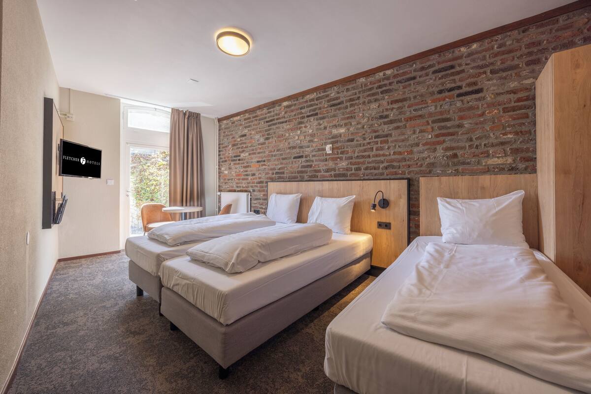 Habitación de hotel con dos camas, una pared de ladrillo, un televisor y un ventanal con cortinas.