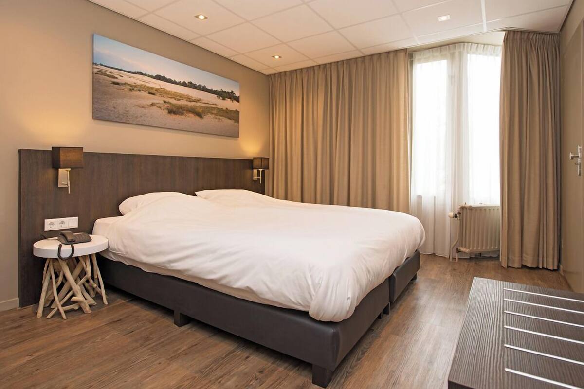 A hotel room with a large bed, a bedside table with a lamp, a painting on the wall, and a view of the outdoors through the window.