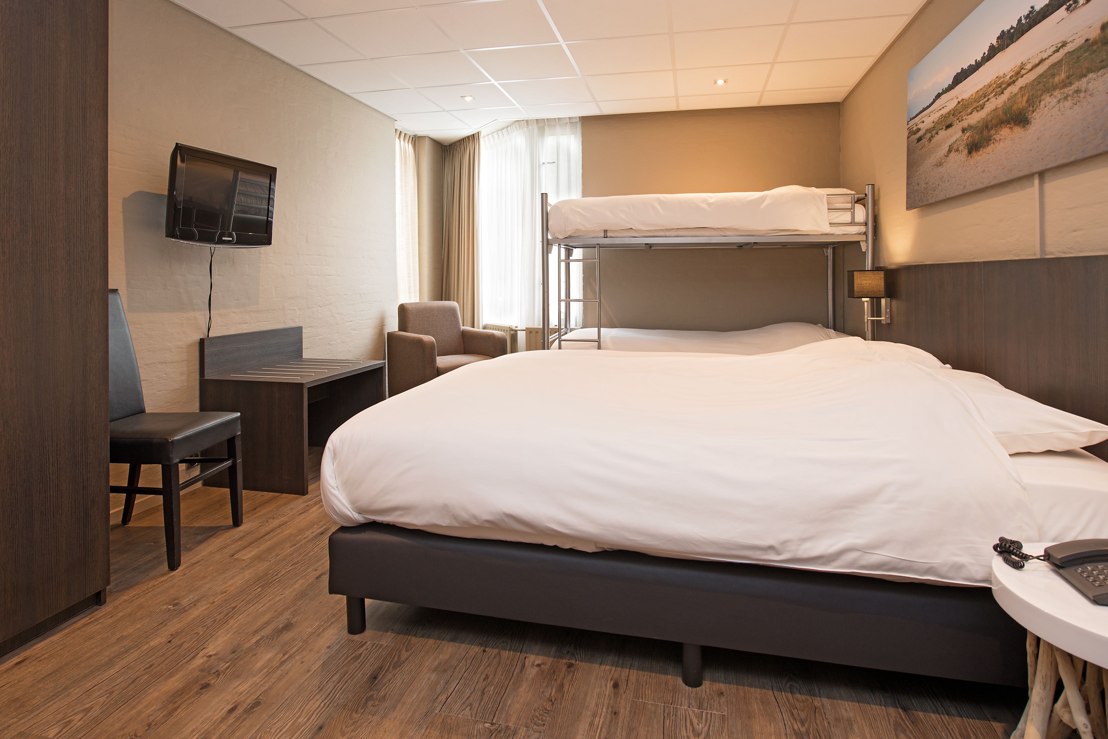 A hotel room with a large bed, a desk with a chair, a TV mounted on the wall, and a bunk bed.