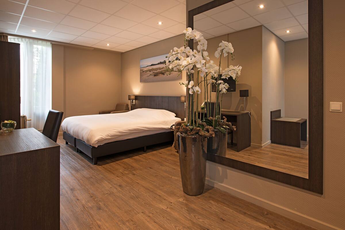 A modern hotel room with a large bed, a desk, a mirror, and a vase with white flowers.