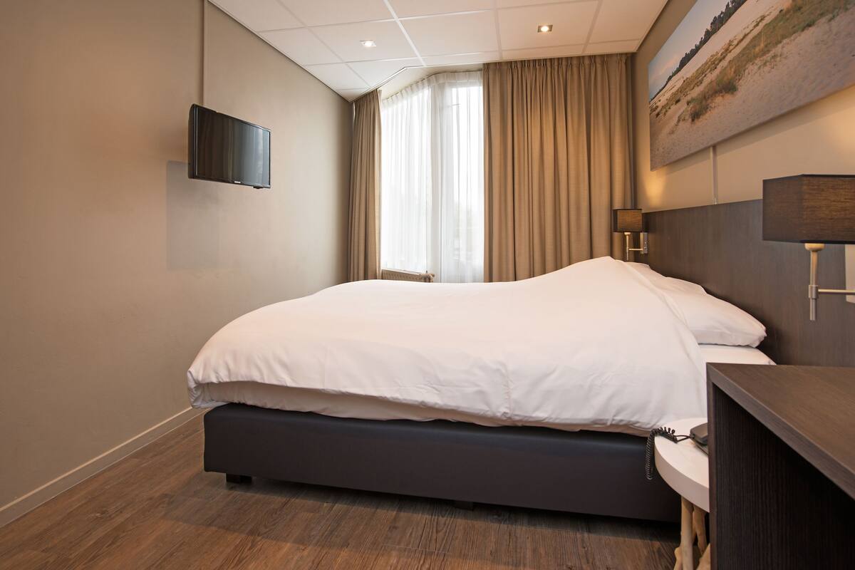 A modern hotel room with a large bed, a flat-screen TV mounted on the wall, a bedside table with a lamp, and a window with curtains.